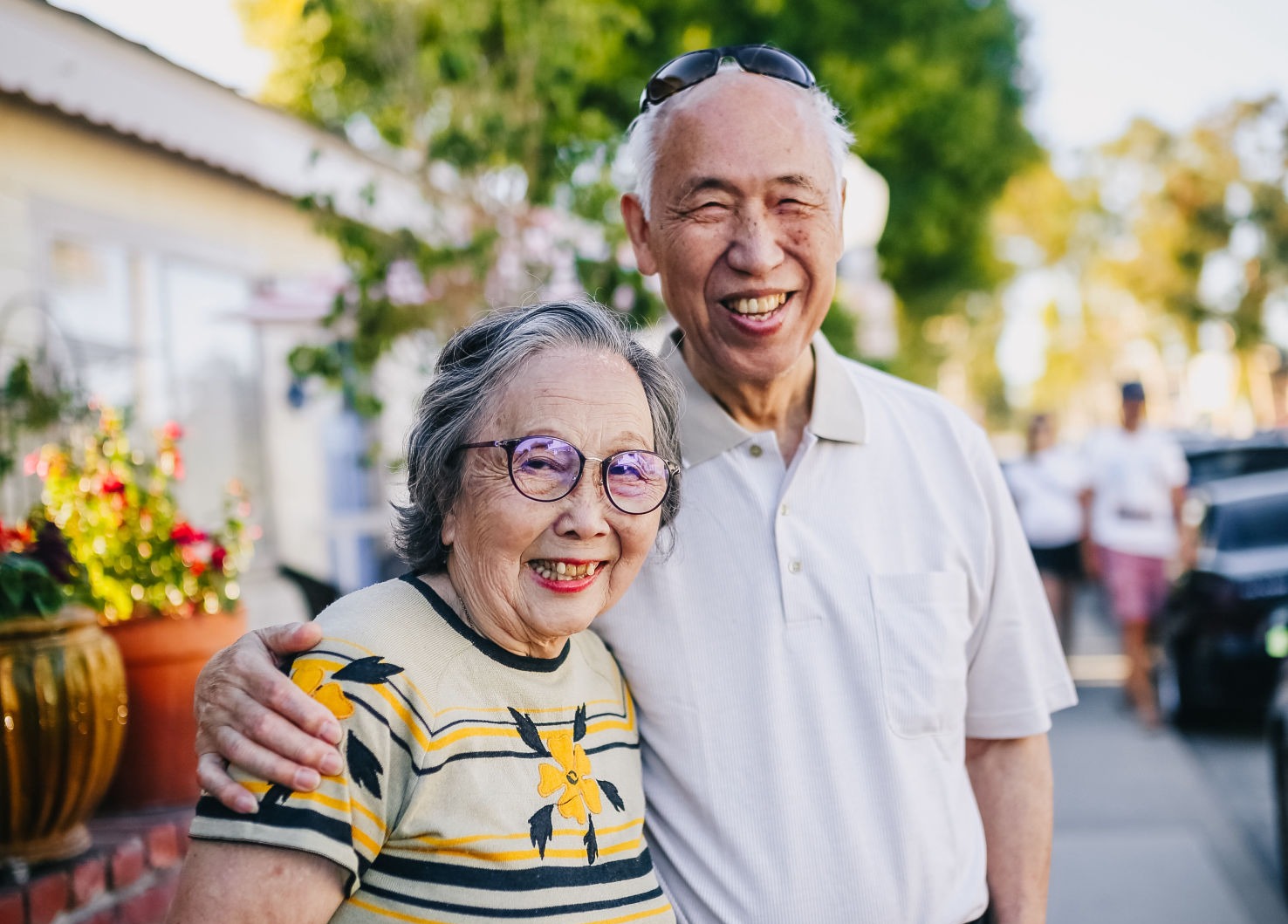 A man and woman smile at the camera. He has his arm around her shoulder. They are standing on a sidewalk, out of focus in the background, on a sunny day