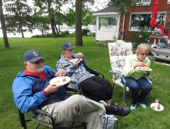Three people sitting on patio chairs on a grass lawn eat off paper plates. A brick house, trees, and a body of water can be seen in the background.
