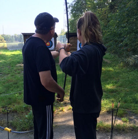 Two people, standing beside each other, hold an archery bow between them. They face away, towards a target that can be seen in the background. The person on the right, with a ponytail, pulls on the bowstring.